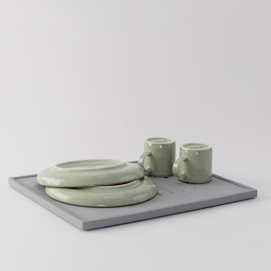 Set of green ceramic items including a bowl, two mugs, and a quick drying dish pad on a light gray background