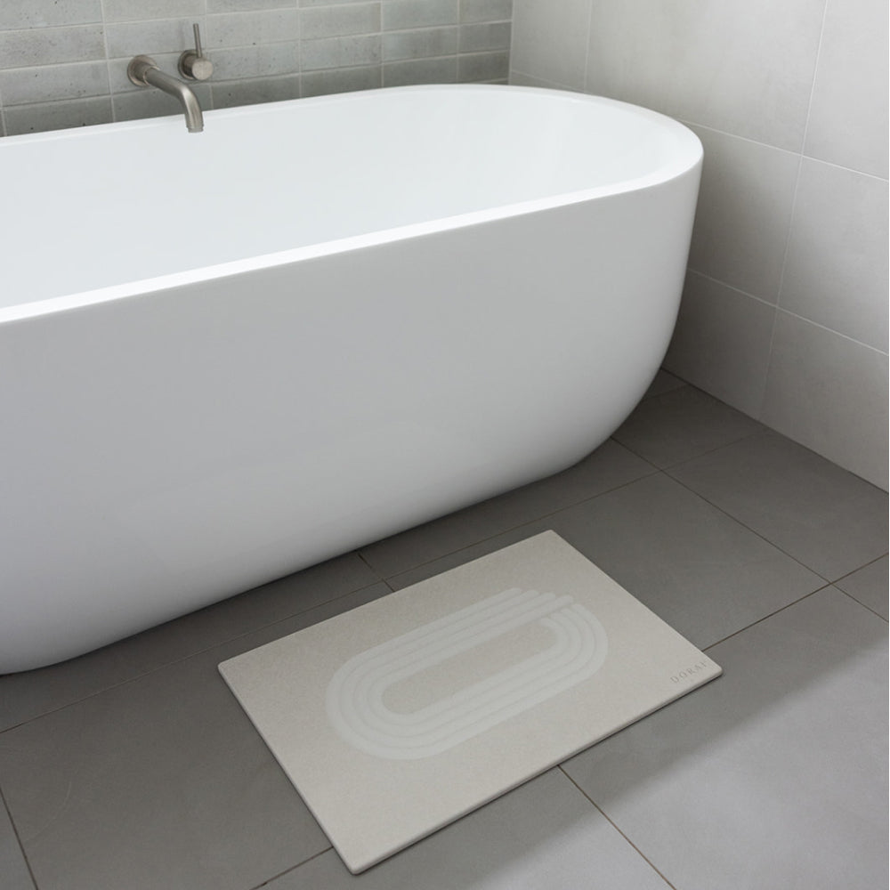 Beige bath mat on a gray tiled bathroom floor next to a white bathtub.