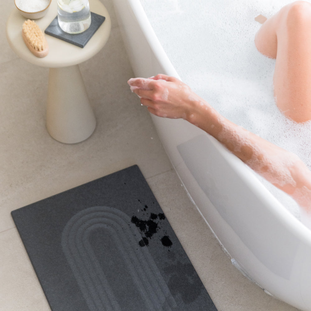 Person taking a bath with a dark grey bath mat on the floor next to the tub.