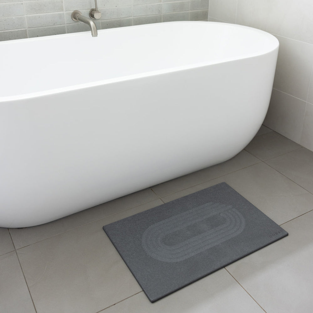 Gray bath mat in front of a white freestanding bathtub on a tiled bathroom floor.