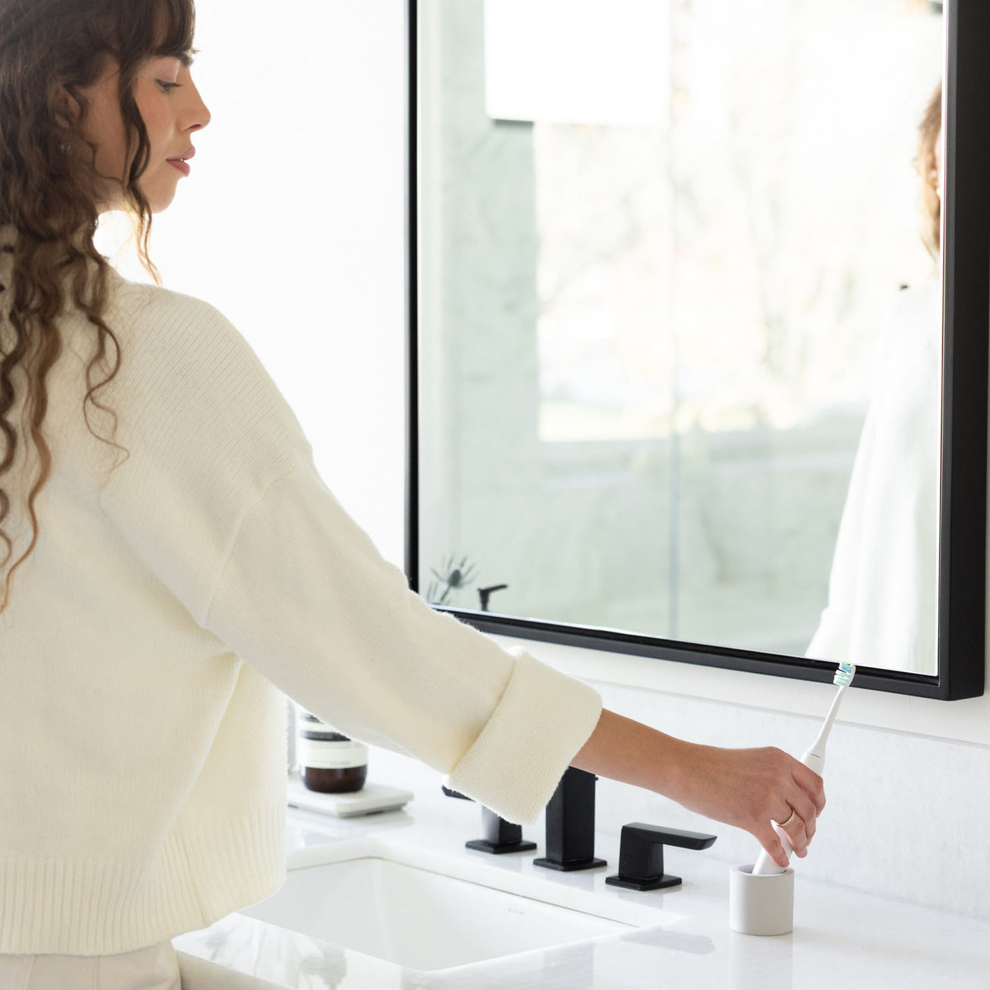 woman taking an electric toothbrush out of a stone electric toothbrush holder with a bathroom setting in the background.