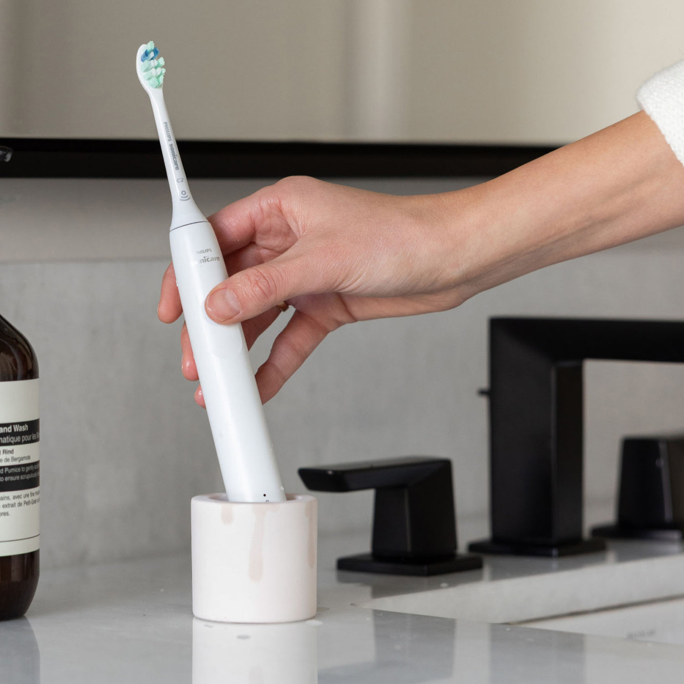 Hand taking an electric toothbrush out of a stone electric toothbrush holder with a bathroom setting in the background.