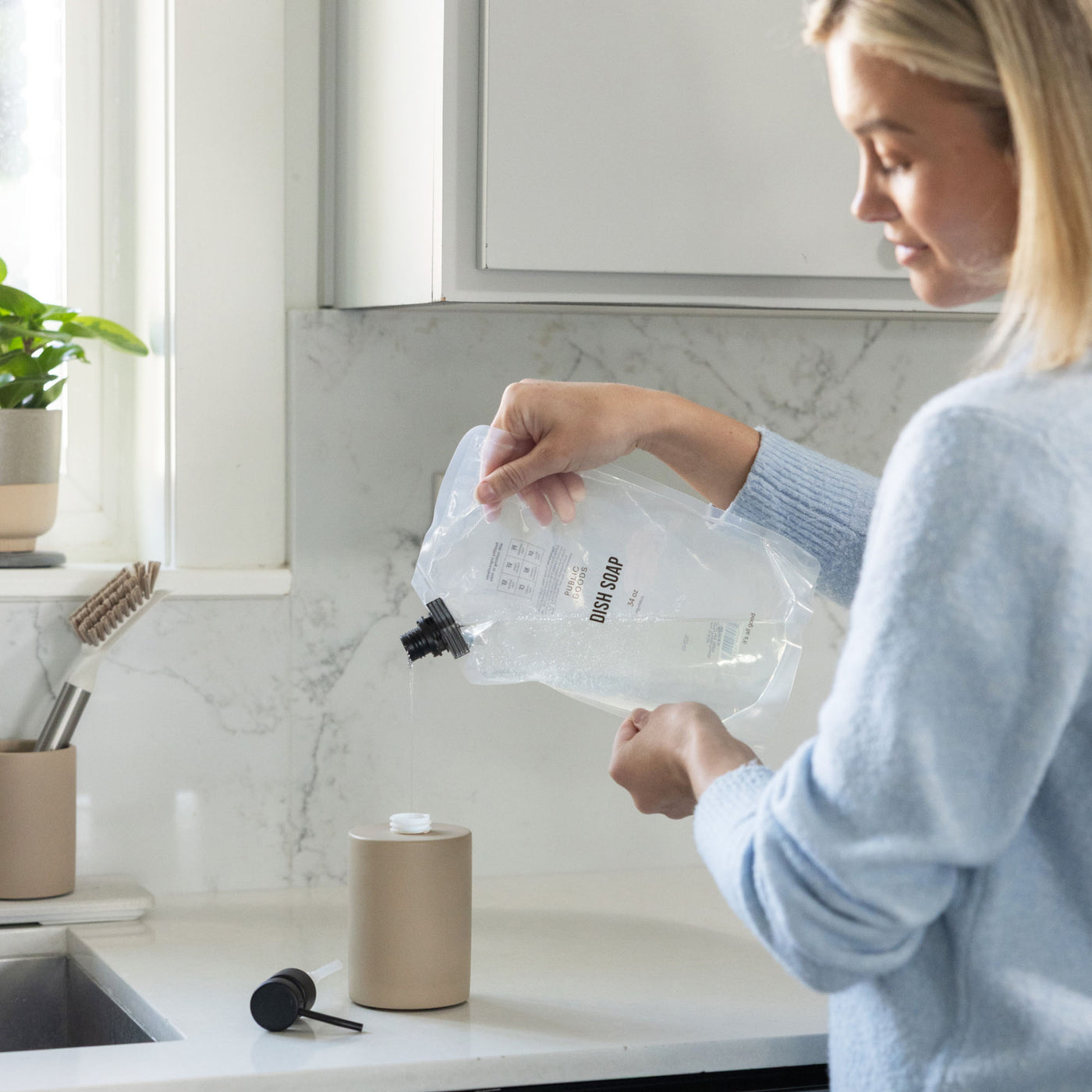 Refilling taupe soap holder with refillable dish soap in kitchen.