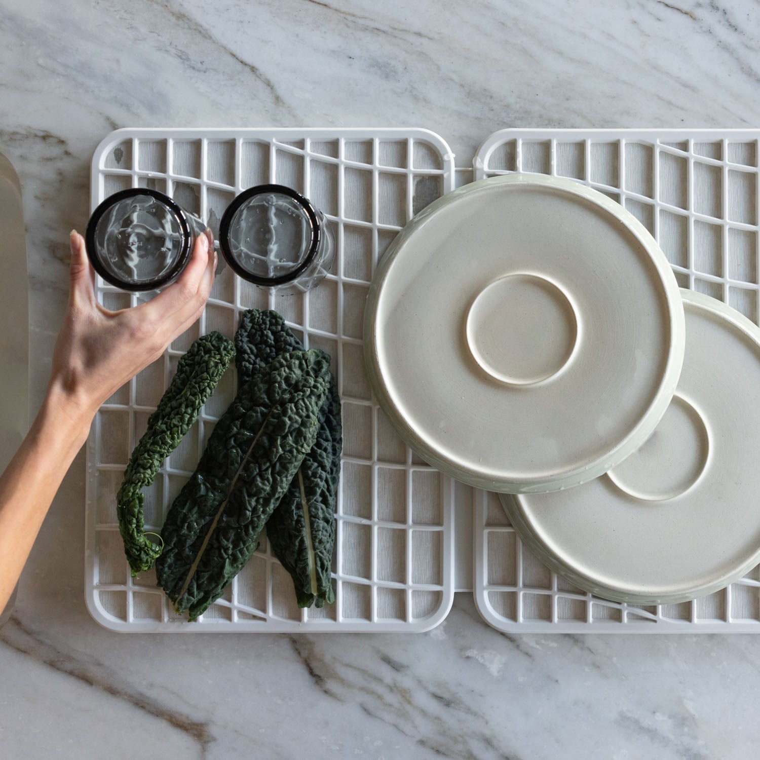 Person holding a glass over a white dish pad with plates and vegetables on a marble countertop.