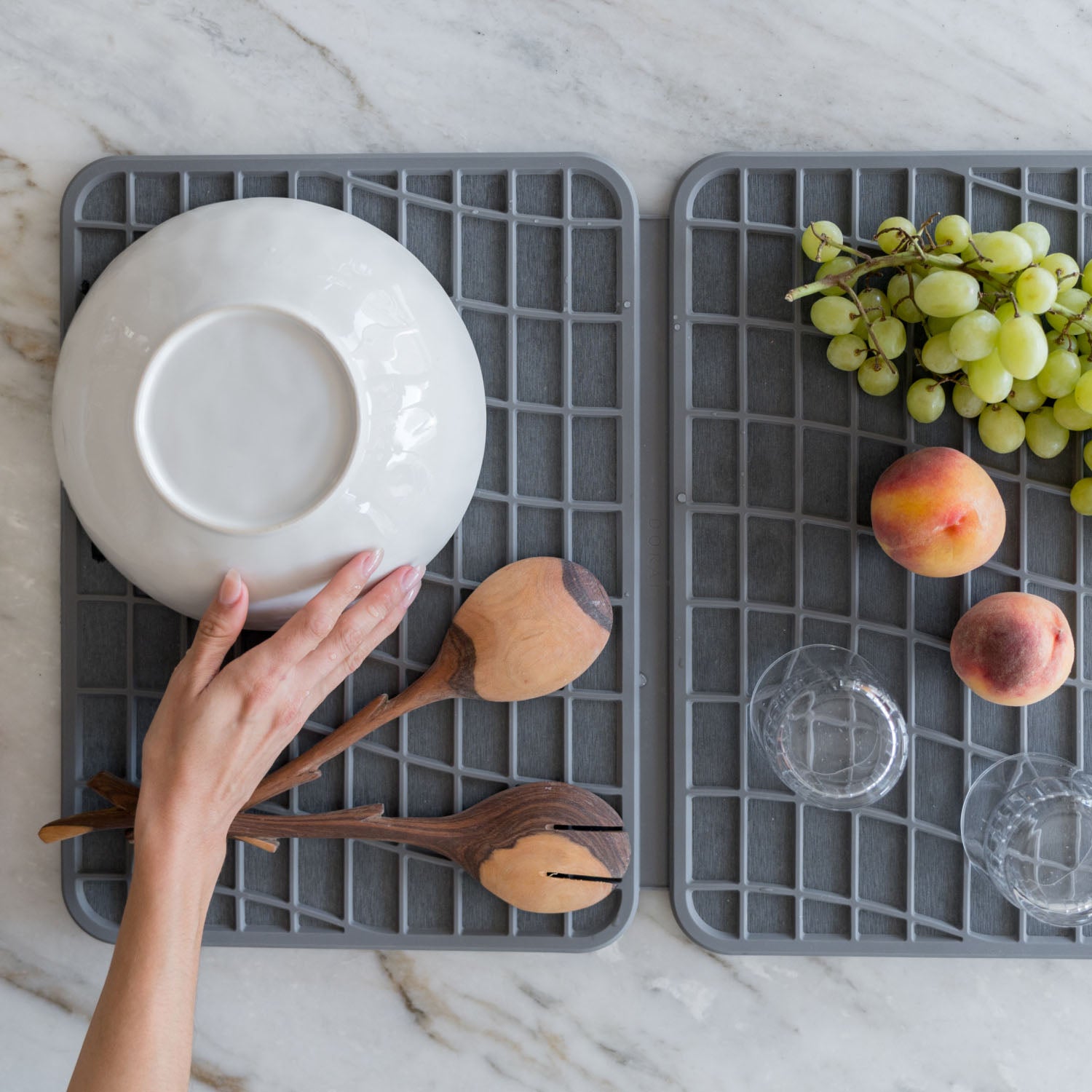 Gray mesh dish drying racks with a white bowl, wooden spoons, grapes, and peaches on a marble countertop.