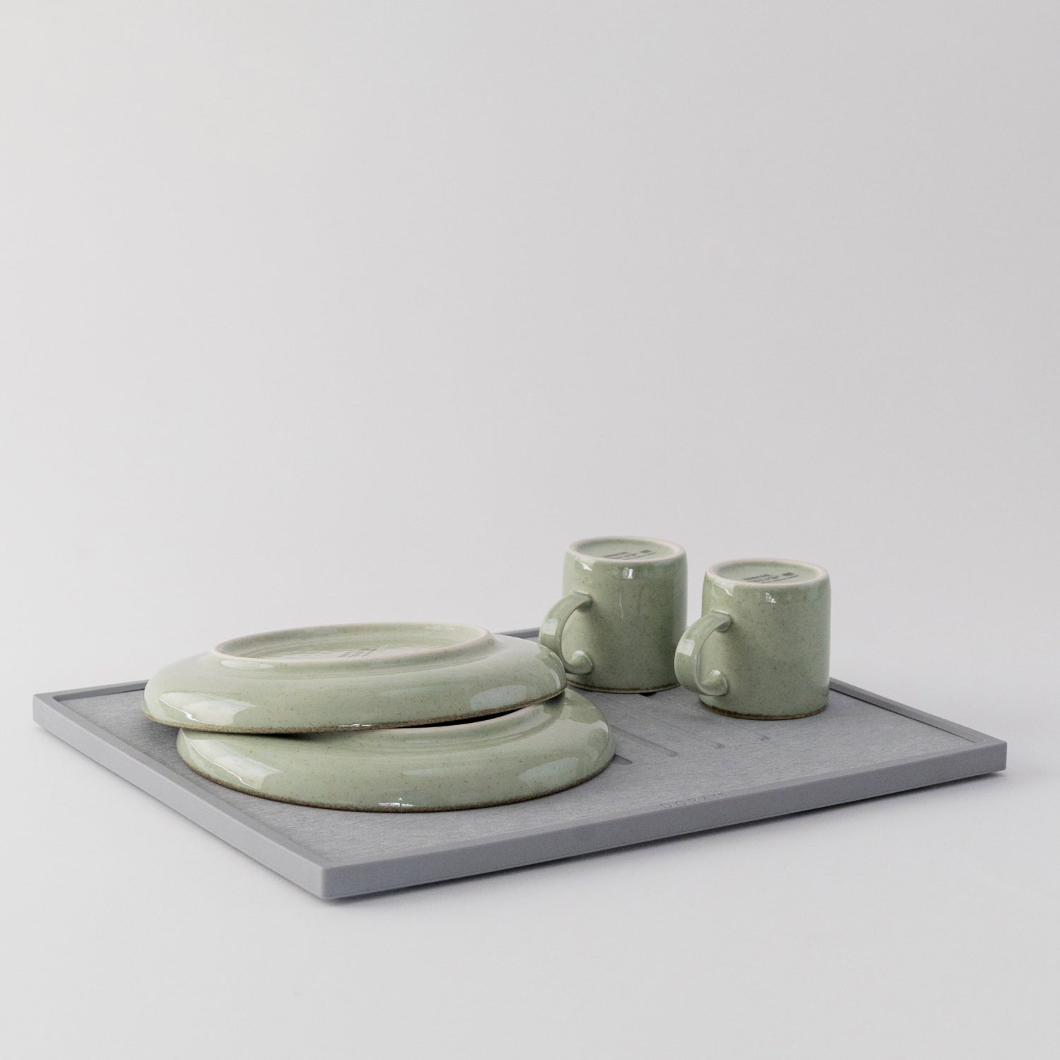 Set of green ceramic items including a bowl, two mugs, and a quick drying dish pad on a light gray background