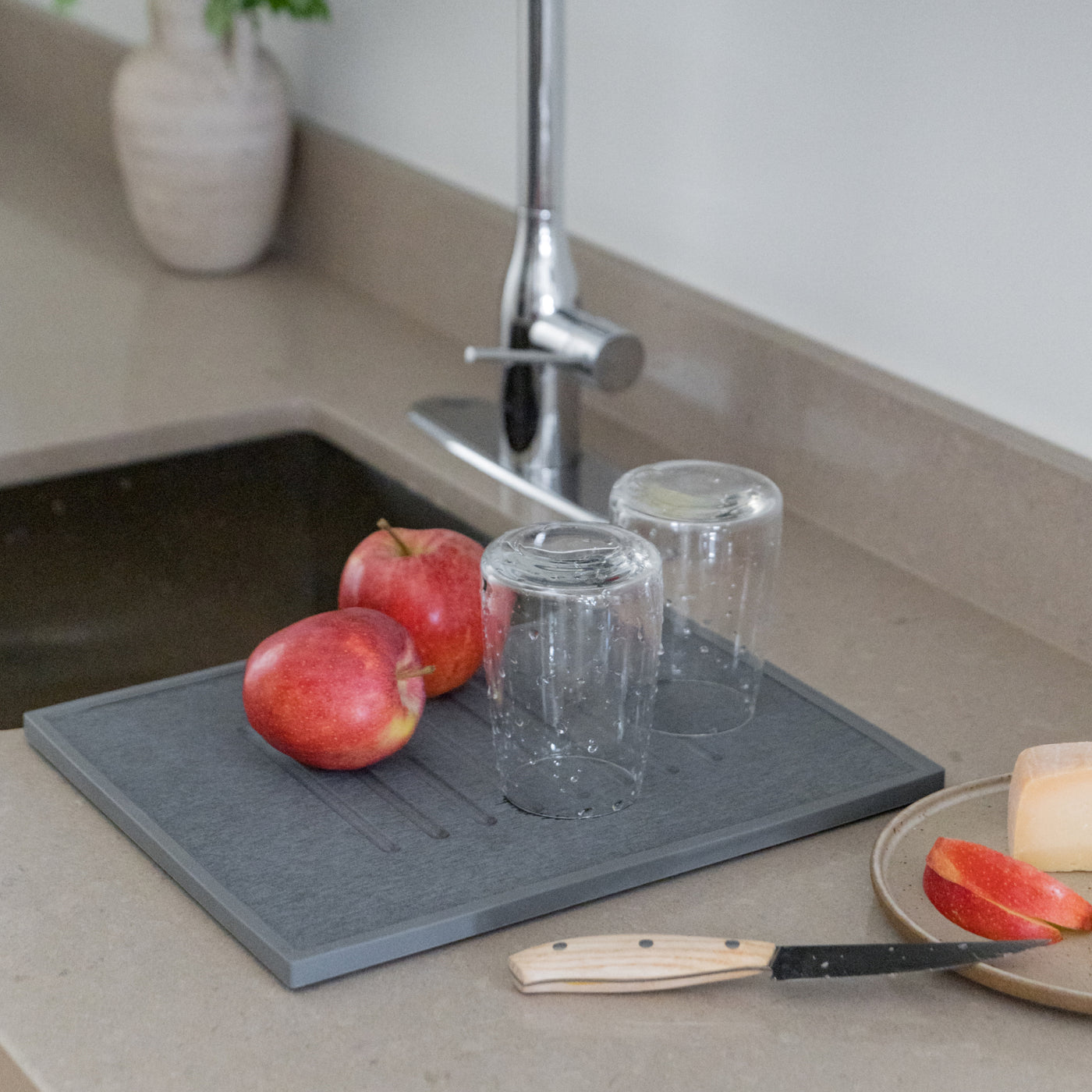 Gray drying mat with apples, glasses, and a knife on a kitchen counter