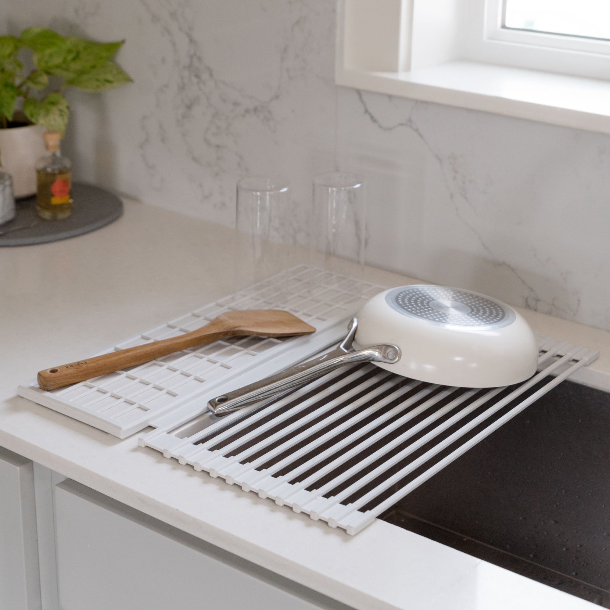Dish pad rolled over a kitchen sink with a white frying pan, wooden spatula, and glass on a marble countertop.